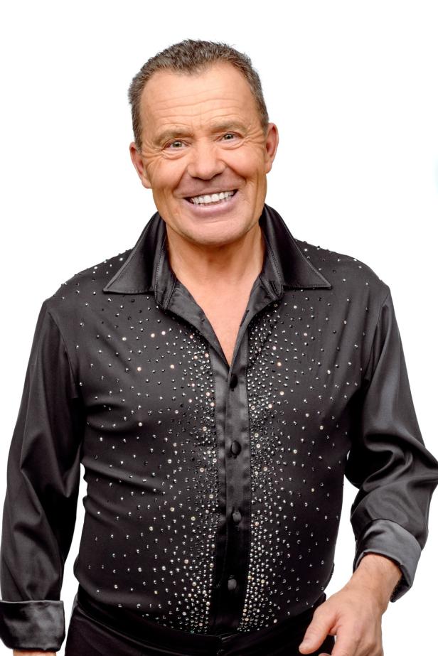 Ein lächelnder Mann in einem schwarzen, glitzernden Hemd vor einem weißen Hintergrund.