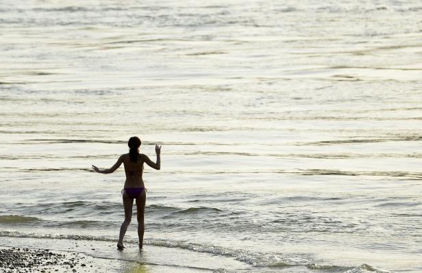Eine Frau steht im Bikini am Ufer und blickt auf das Meer.