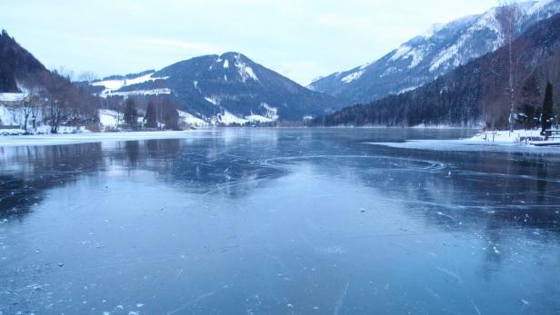 Ein zugefrorener See liegt vor einer verschneiten Berglandschaft.
