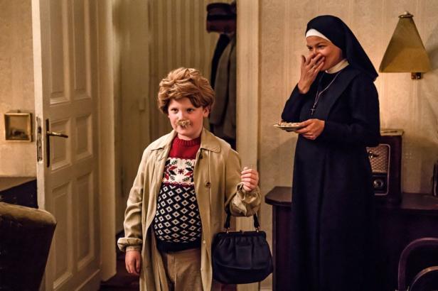 Ein Junge mit aufgeklebtem Schnurrbart und eine Nonne in einem Innenraum.