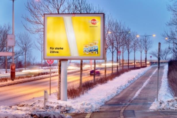 Eine Litfaßsäule mit Werbung für Zahnpasta steht an einer verschneiten Straße.