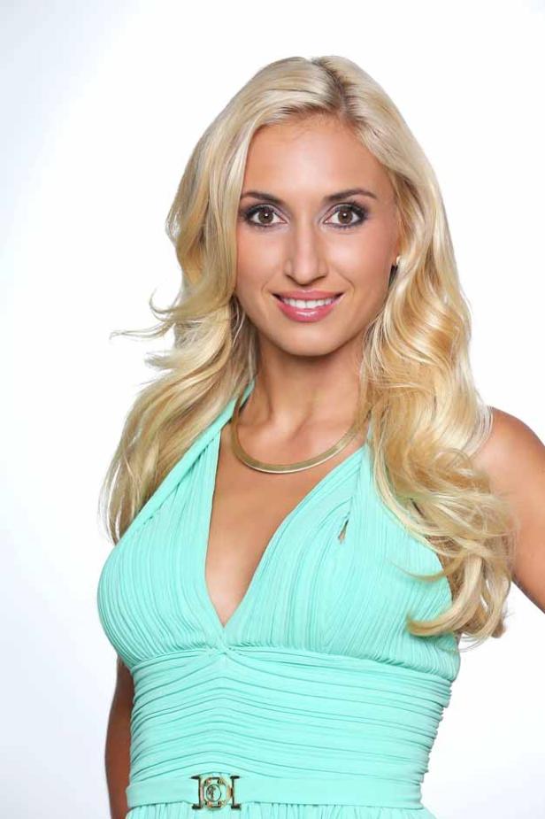 Eine blonde Frau lächelt in einem türkisfarbenen Kleid mit goldenem Schmuck.