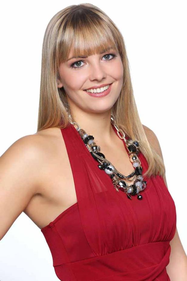 Eine lächelnde Frau mit blonden Haaren und einem roten Kleid mit einer auffälligen Halskette.