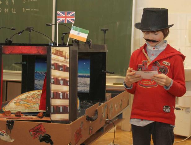 Ein Junge mit aufgeklebtem Schnurrbart liest vor einem selbstgebauten Schiffsmodell mit Flaggen von Großbritannien und Indien.