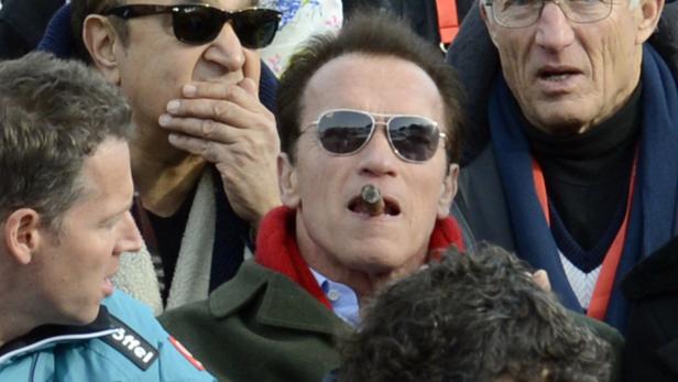 Arnold Schwarzenegger raucht eine Zigarre, umgeben von anderen Zuschauern.