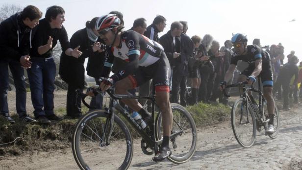 Zwei Radrennfahrer fahren auf einem holprigen Kopfsteinpflasterweg, während Zuschauer am Straßenrand stehen.
