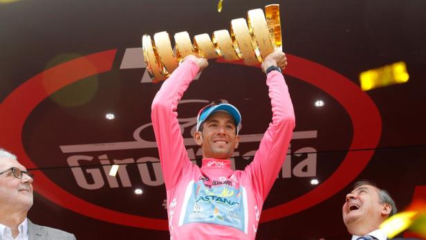 Ein Radrennfahrer im rosa Trikot stemmt eine goldene Trophäe in die Höhe.