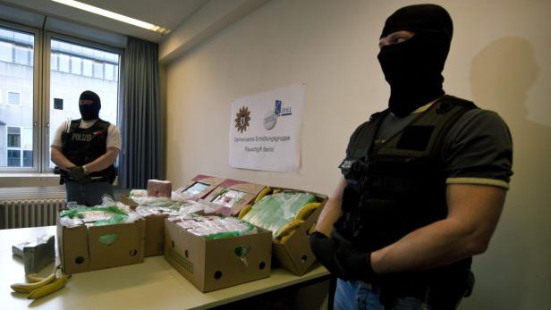 Zwei maskierte Zollbeamte stehen neben beschlagnahmten Drogen in Kartons.
