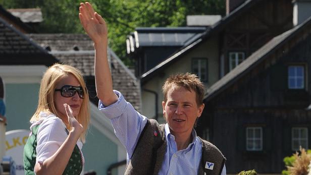 Zwei Personen winken vor dem Hintergrund von Häusern.