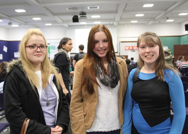 Drei junge Frauen stehen lächelnd in einem Raum mit Publikum im Hintergrund.