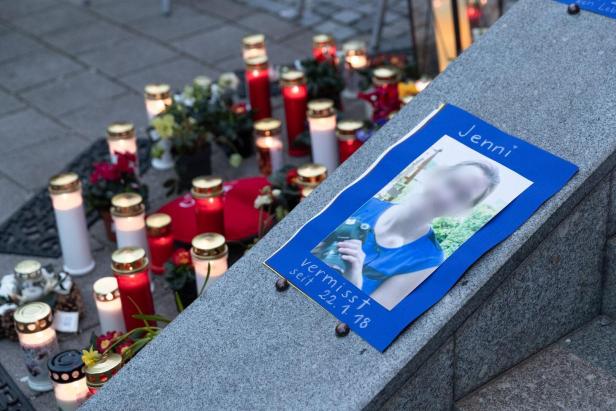 Ein Suchplakat für die vermisste Jenni liegt auf einer Steinmauer, umgeben von zahlreichen brennenden Grablichtern und Blumen.