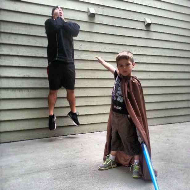 Ein Junge im Umhang hält ein Spielzeug-Lichtschwert, während ein Mann im Hintergrund springt.