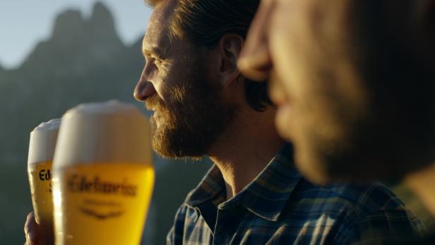 Edelweiss Weizen Bier TV-Spot 2 Zwei Männer stoßen mit Gläsern Edelweiss-Bier vor einer Bergkulisse an.