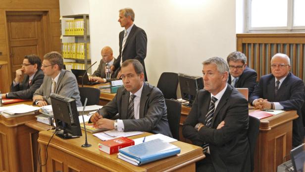 Mehrere Männer in Anzügen sitzen an Tischen in einem Gerichtssaal.