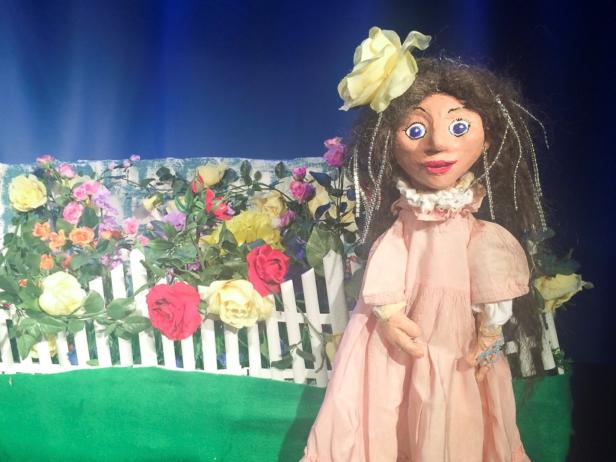 Eine Puppe mit einem rosa Kleid steht vor einem weißen Zaun mit Blumen.