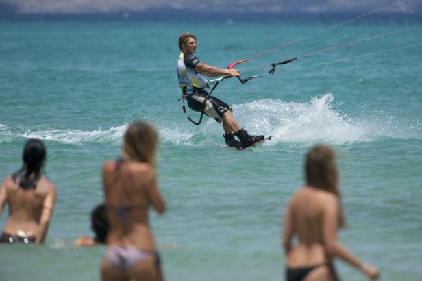 Ein Kitesurfer fährt auf dem Wasser, während Badegäste im Vordergrund stehen.