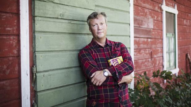 Ein Mann im karierten Hemd lehnt an einer Holzwand.