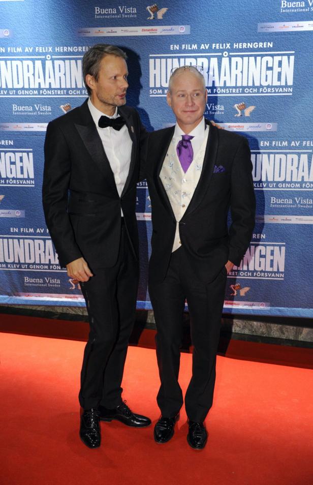 Zwei Männer in Anzügen posieren vor einem Filmplakat für „Hundraåringen som klev ut genom fönstret och försvann“.