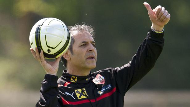 Ein Mann im Trainingsanzug hält einen Fußball in der Hand und zeigt den Daumen nach oben.