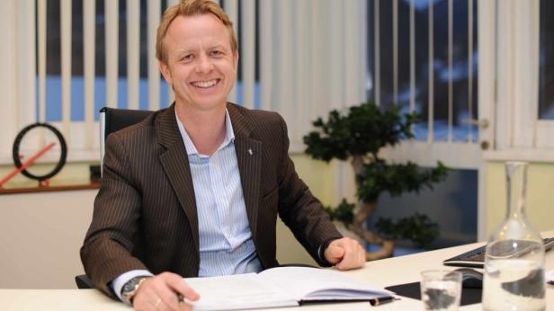 Ein lächelnder Mann im Anzug sitzt an einem Schreibtisch mit einem offenen Buch.