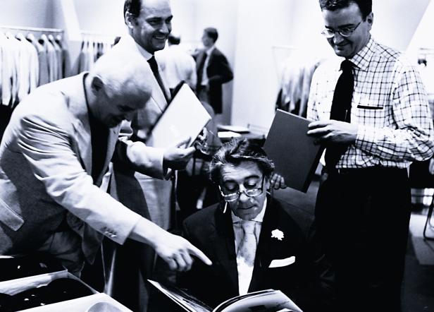 Ein Mann mit Brille sitzt und schaut ein Buch an, umgeben von drei anderen Männern in einem Bekleidungsgeschäft.