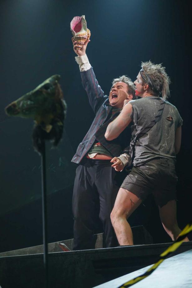 Zwei Schauspieler auf einer Bühne, einer hält eine Muschel hoch, während der andere ihn festhält.