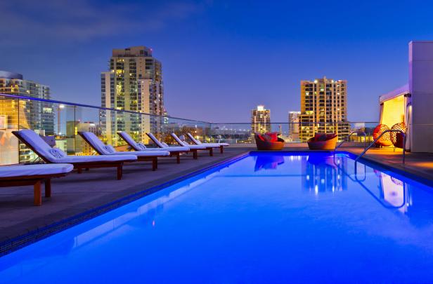 Ein beleuchteter Pool auf einem Dach mit Liegestühlen und Blick auf die Skyline der Stadt bei Nacht.