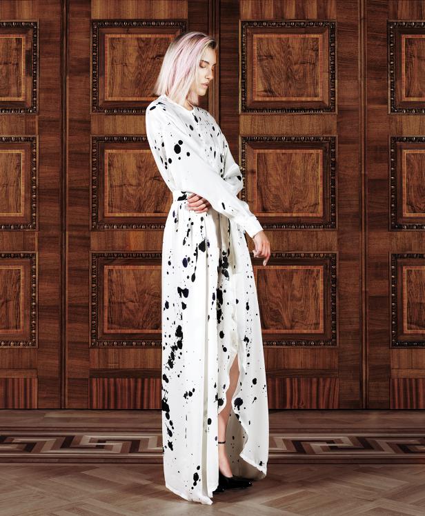 Eine Frau mit rosa Haaren trägt ein weißes Kleid mit schwarzen Farbspritzern vor einer Holzwand.