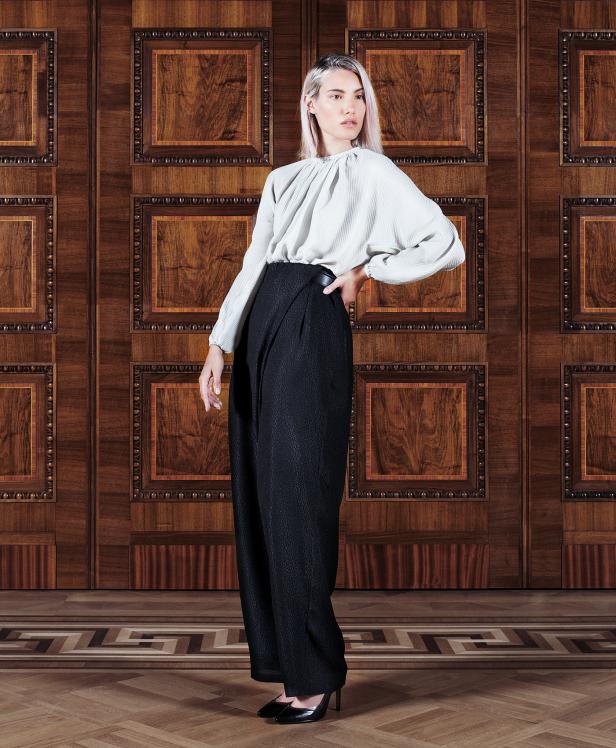 Eine Frau posiert in einer hellen Bluse und einer weiten, schwarzen Hose vor einer verzierten Holzwand.