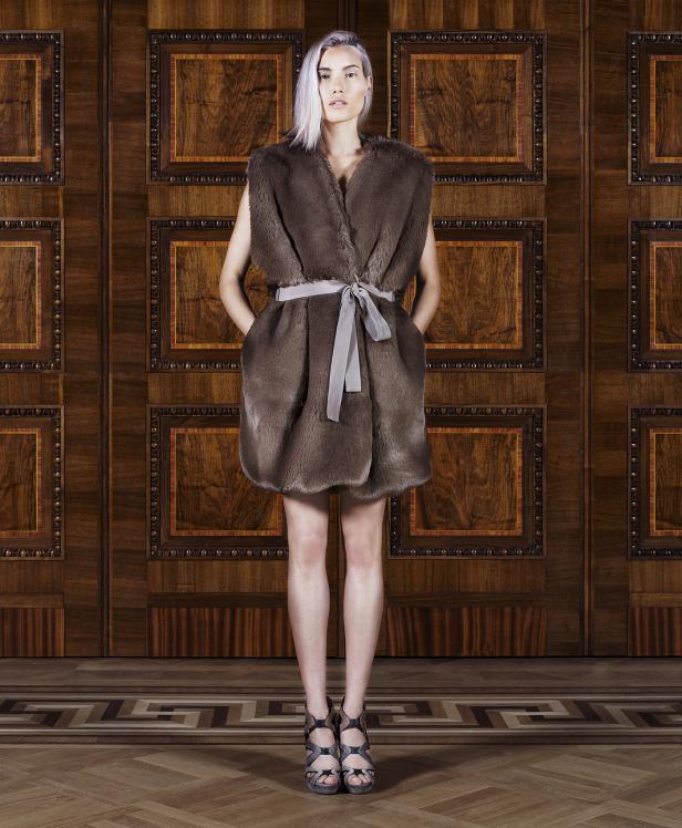 Eine Frau mit hellviolettem Haar trägt einen braunen Pelzmantel mit grauem Gürtel vor einer Holzwand.