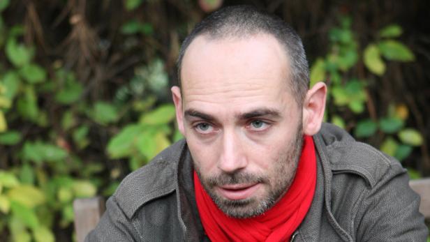 Ein Mann mit rotem Schal und grauem Jackett sitzt draußen vor grünem Laub.