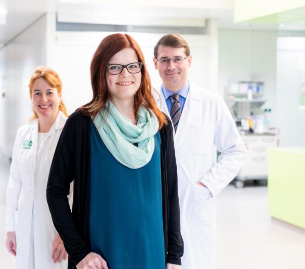 Eine junge Frau mit Brille steht vor zwei Ärzten in einem hellen Flur.