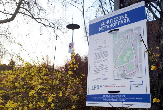 Ein Schild mit der Aufschrift „Schutzzone Metahofpark“ steht neben gelb blühenden Büschen.