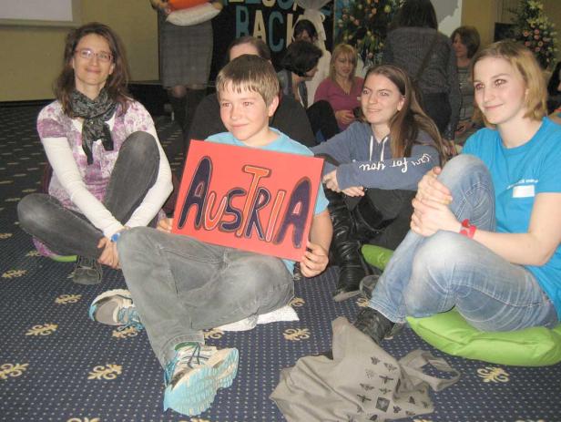 Eine Gruppe junger Leute sitzt auf dem Boden, ein Junge hält ein Schild mit der Aufschrift „Austria“.