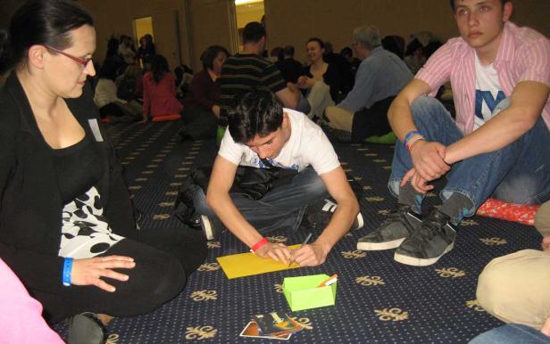 Mehrere Personen sitzen auf dem Teppich, ein junger Mann schreibt auf ein gelbes Blatt Papier.