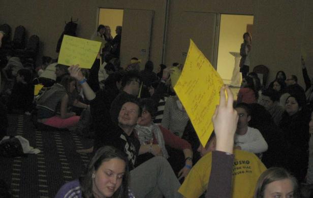 Eine Gruppe von Menschen sitzt auf dem Boden und hält gelbe Schilder hoch.