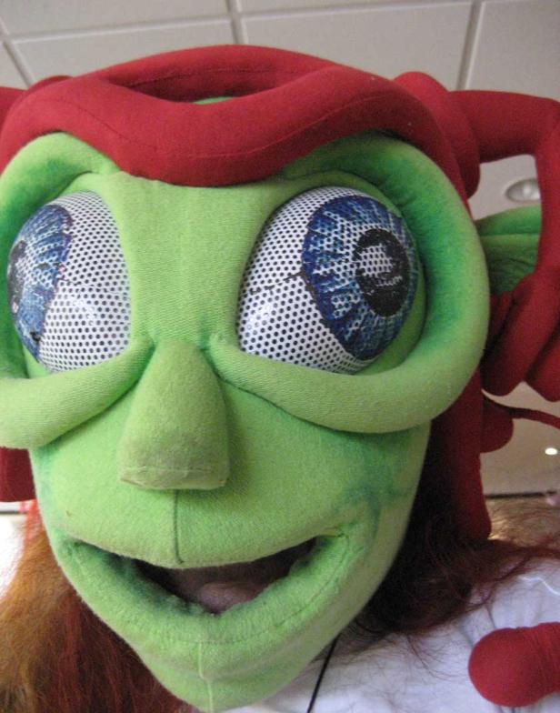 Nahaufnahme einer Person, die eine grüne Alien-Maske mit großen Augen und rotem Kopfschmuck trägt.