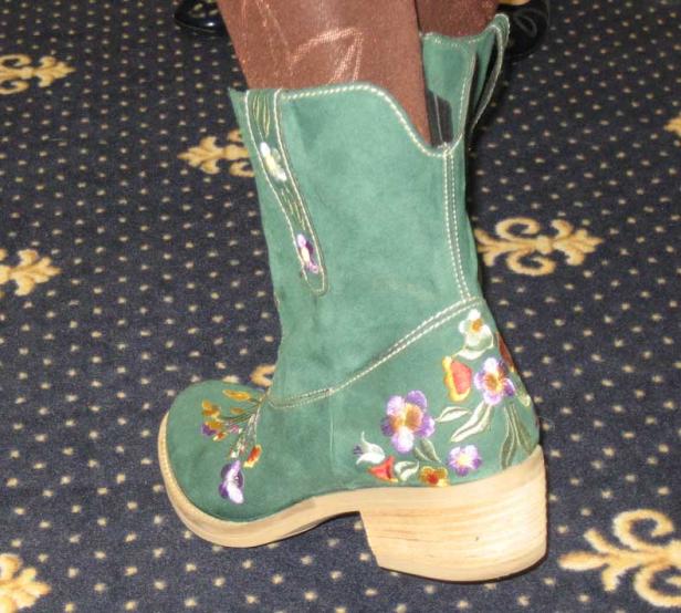 Ein grüner Wildlederstiefel mit Blumenstickereien steht auf einem gemusterten Teppich.