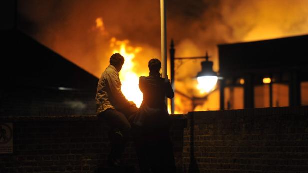 BRITAIN RIOTS Zwei Personen beobachten nachts ein Feuer hinter einer Mauer.