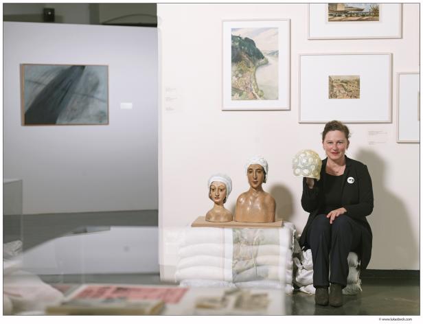 Eine Frau sitzt in einer Galerie neben zwei Büsten und hält eine Badekappe hoch.