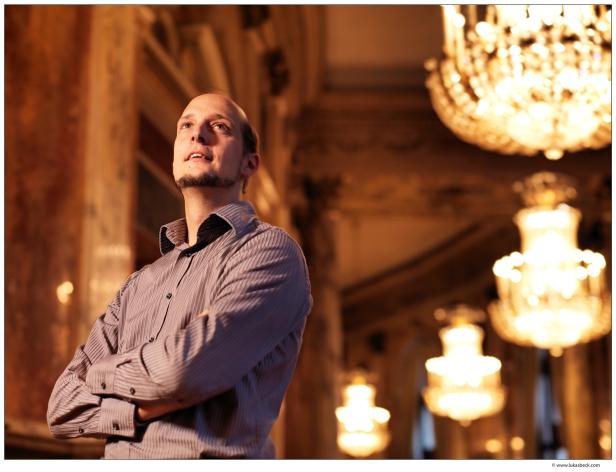 Ein Mann steht mit verschränkten Armen in einem eleganten Saal mit Kristalllüstern.