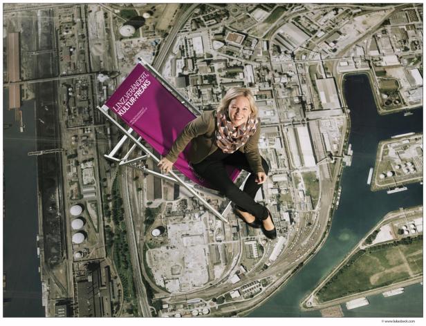 Eine Frau sitzt auf einem lila Liegestuhl mit dem Schriftzug „Linz verändert, Kultur-Freaks“ vor einem Luftbild von Linz.