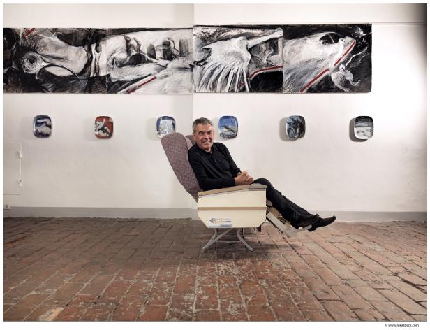 Ein Mann sitzt in einem Flugzeugsitz vor Kunstwerken an einer Wand.