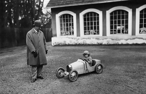 Ein Mann mit Melone beobachtet ein Kind in einem Spielzeugauto.
