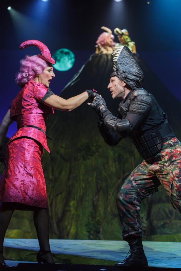 Dinosaurier, Das Musical Zwei Schauspieler in extravaganten Kostümen stehen auf einer Bühne vor einer gemalten Landschaft.