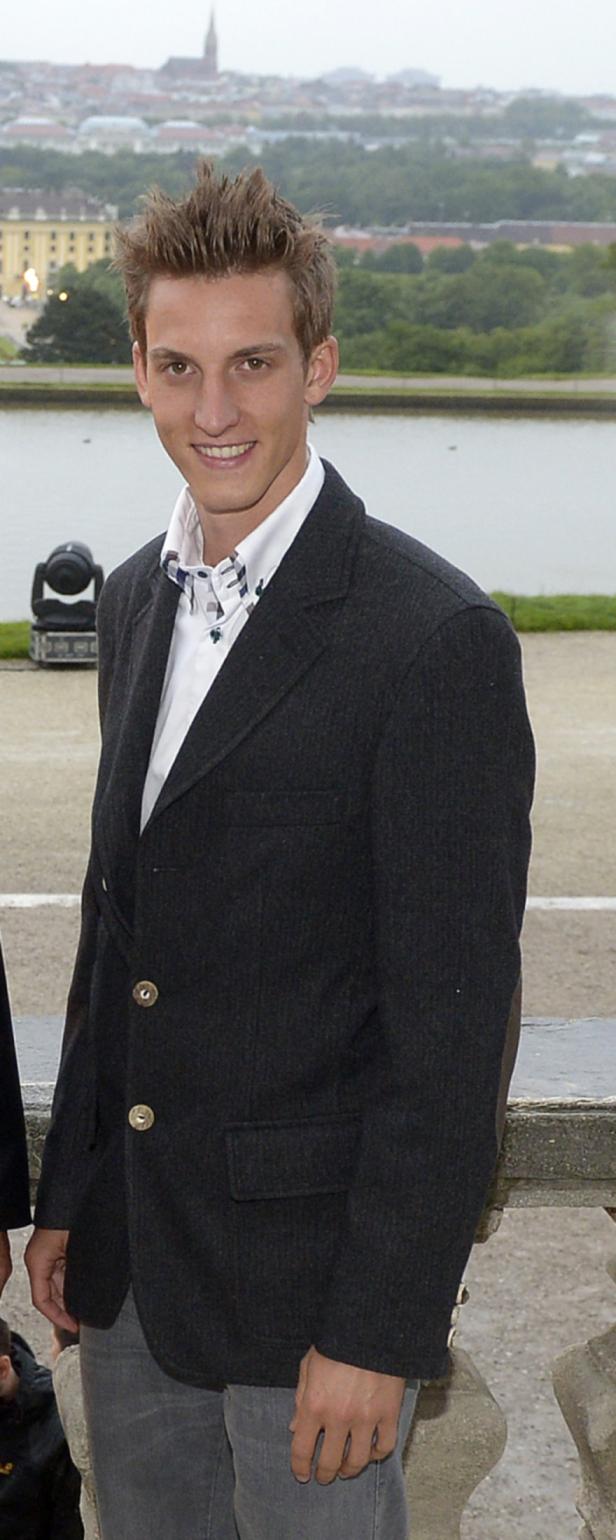 Ein junger Mann in Jackett und Hemd vor einer Parkanlage mit See.