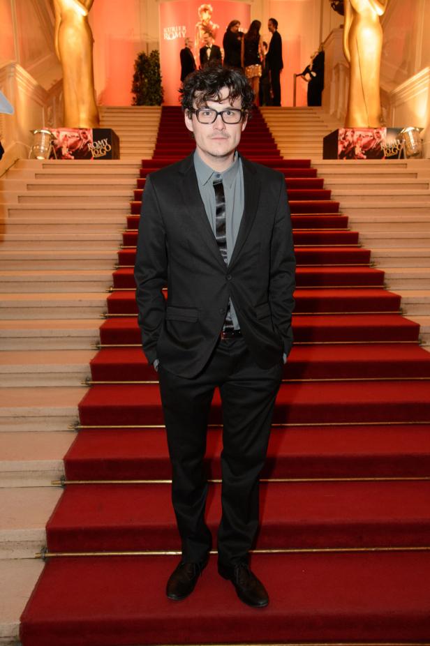 Ein Mann mit Brille steht auf einem roten Teppich vor einer Treppe.