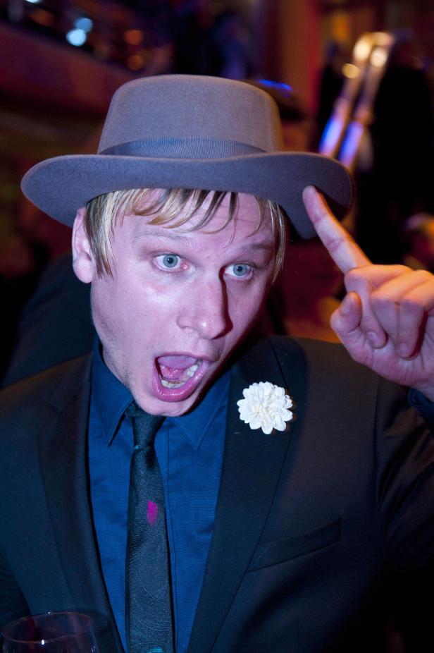 Ein Mann mit Hut, offenem Mund und Blume am Revers blickt in die Kamera.