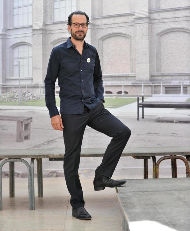 Ein Mann mit Brille posiert mit einem Fuß auf einer Bank vor einem Gebäude.