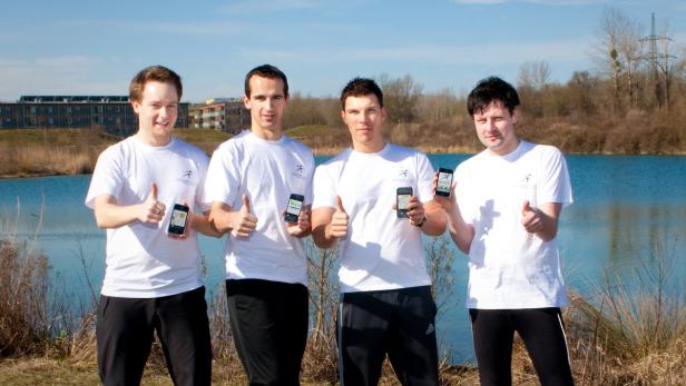 Vier Männer in Sportkleidung präsentieren eine App vor einem See und geben Daumen hoch.